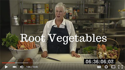 Preview of the Root Vegetables video: a woman wearing a black apron standing behind a cutting board with a knife in her hand