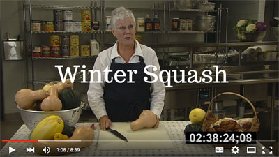 Preview of the Winter Squash video featuring a woman in a black apron standing behind a cutting board and holding a knife and a winter squash