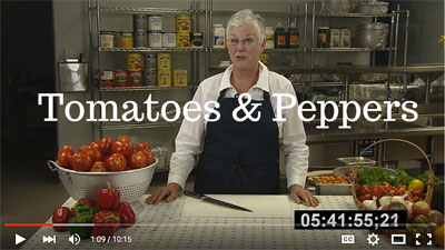 Preview of the tomatoes and peppers video featuring a woman in a black apron standing in front of a cutting board with a variety of tomatoes and peppers around it