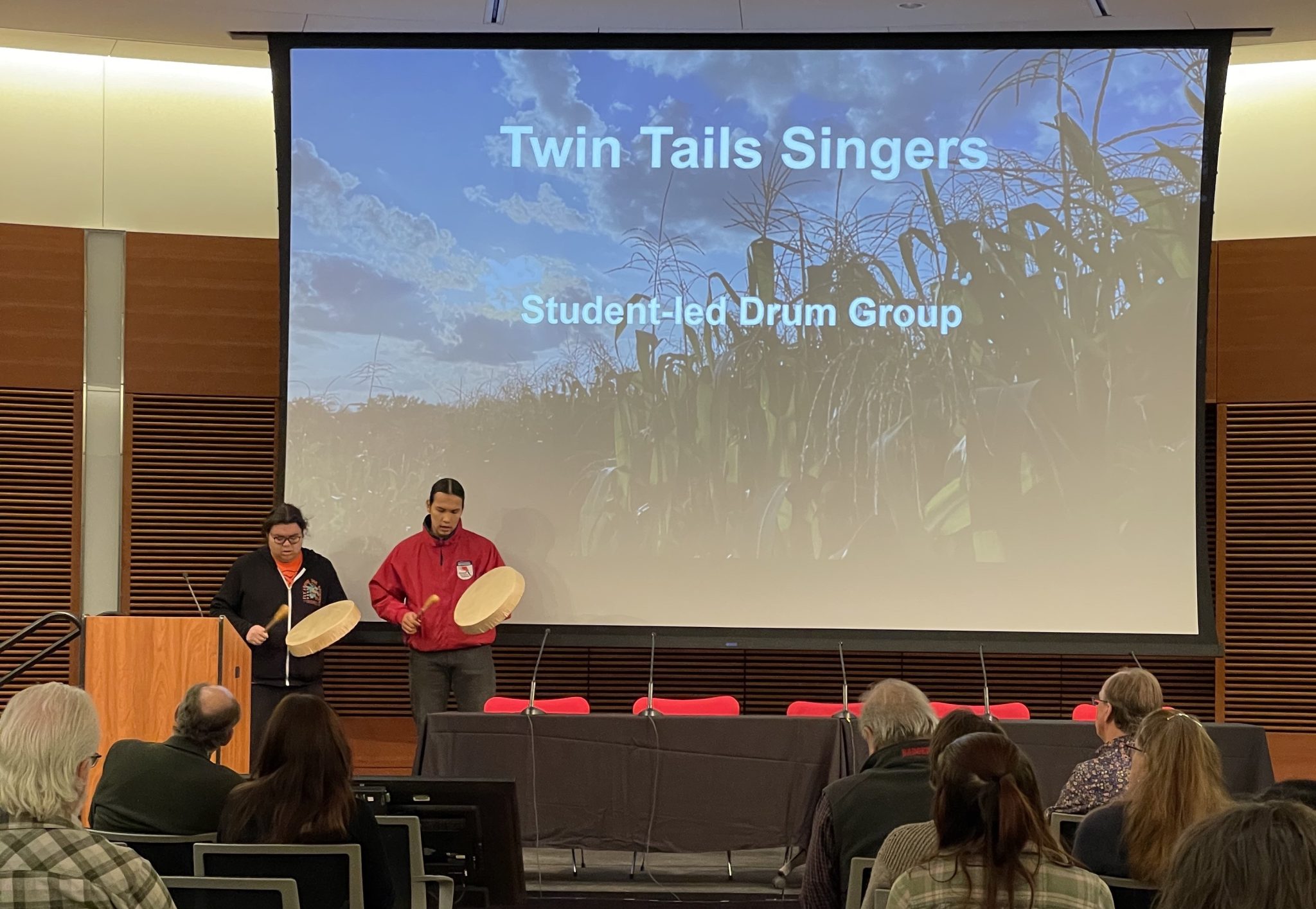 Two people playing a hand held drum in front of a projector screen that says twin tails singers on it
