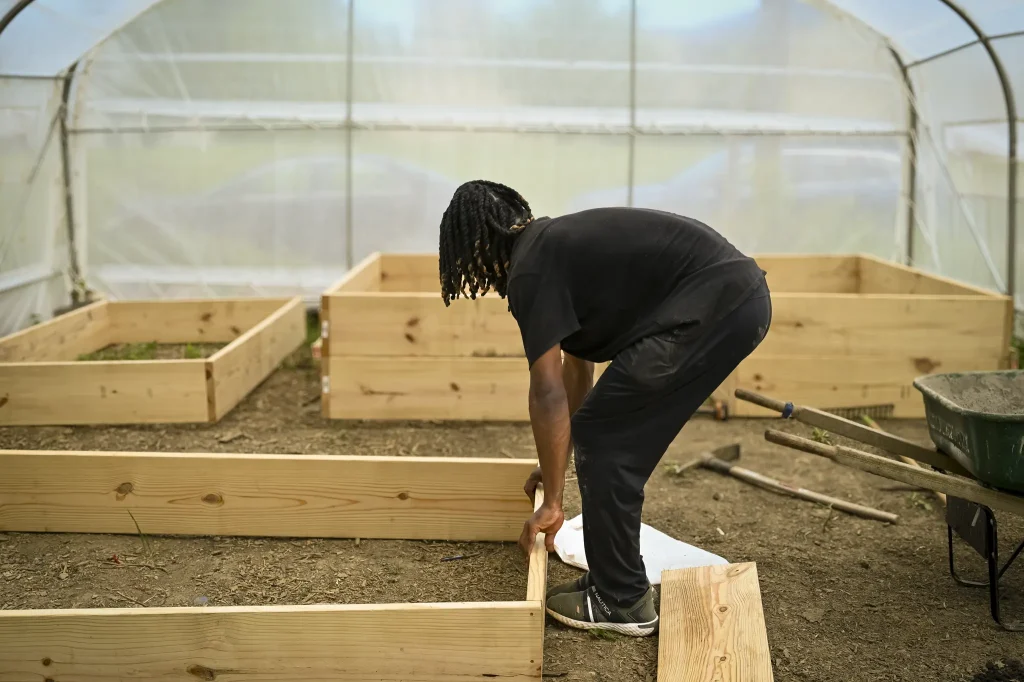 An urban farmer assembles wooden garden beds inside a greenhouse, surrounded by tools, planks, and completed raised beds on a Milwaukee urban farm.