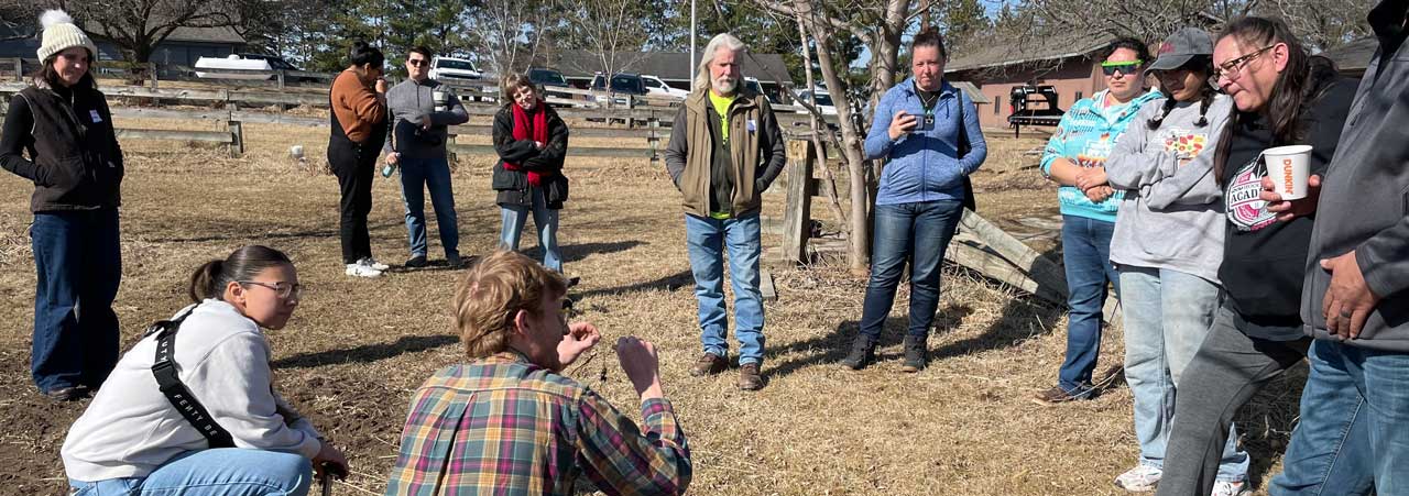 A group of twelve people in a loose circle listening to a person speaking to them in a small pasture