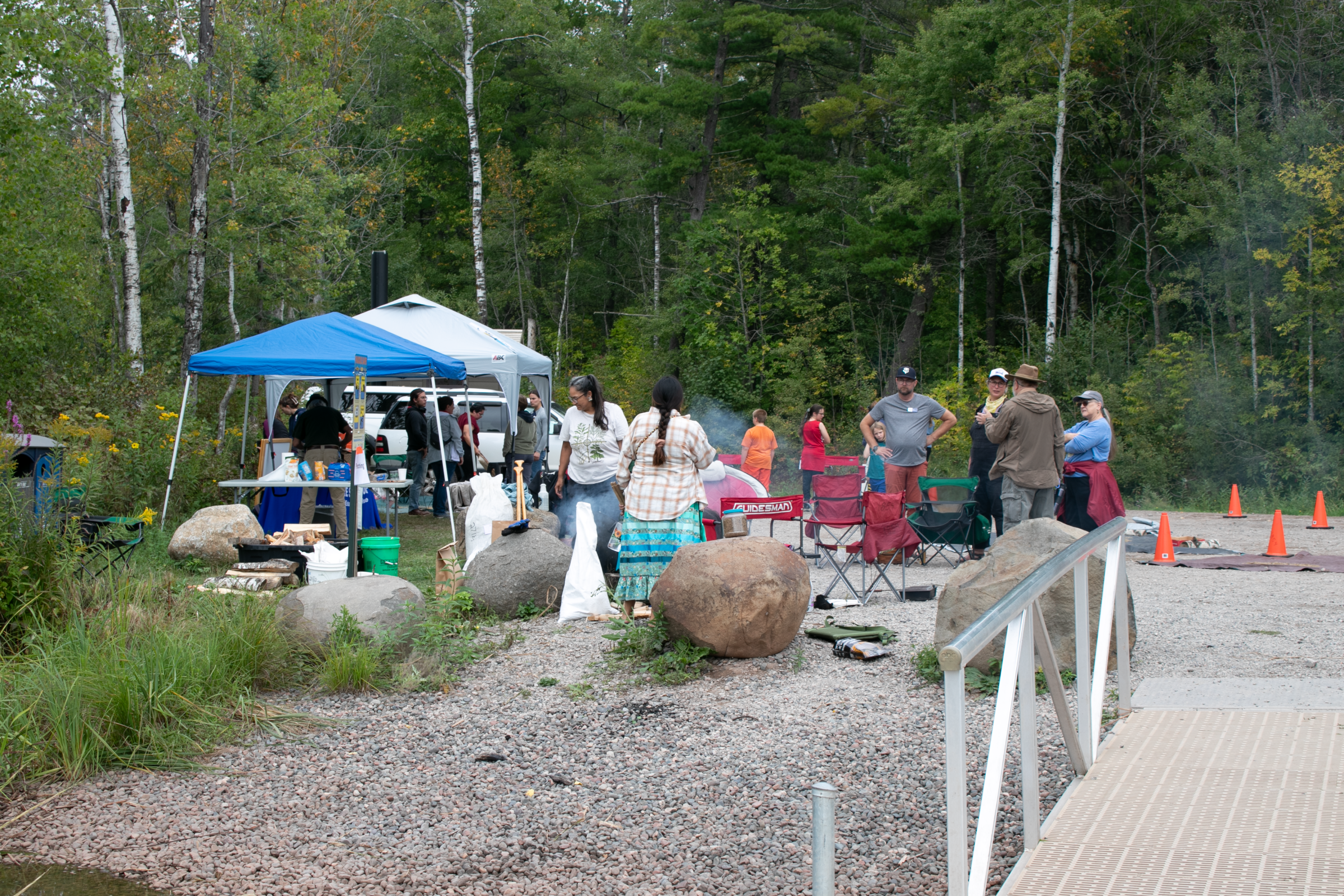 A group of participants at the St. Louis River Estuary Manoomin Camp standing at different wild rice processing stations. A dock is in the foreground and a forest is in the background.