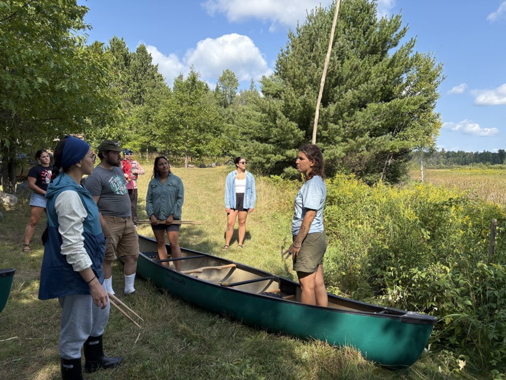 Intertribal Manoomin Camp organizer Sagen Quale stands in a canoe with a tall wooden pole demonstrating to participants how to harvest wild rice and steer the canoe.