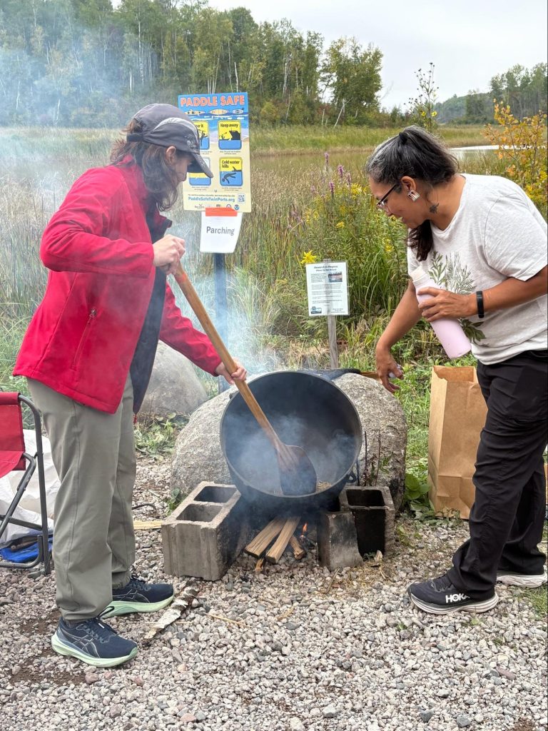 Two people parching wild rice in a kettle over a fire with the St. Louis River in the background.