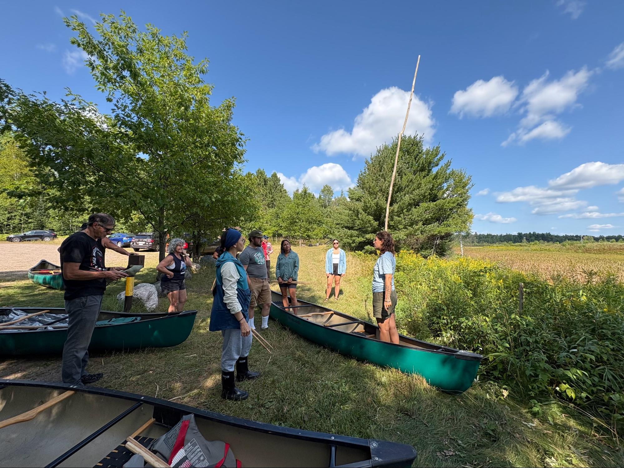 A woman standing in a canoe on the shore holding a long wooden pole and speaking to a group of people also with canoes.