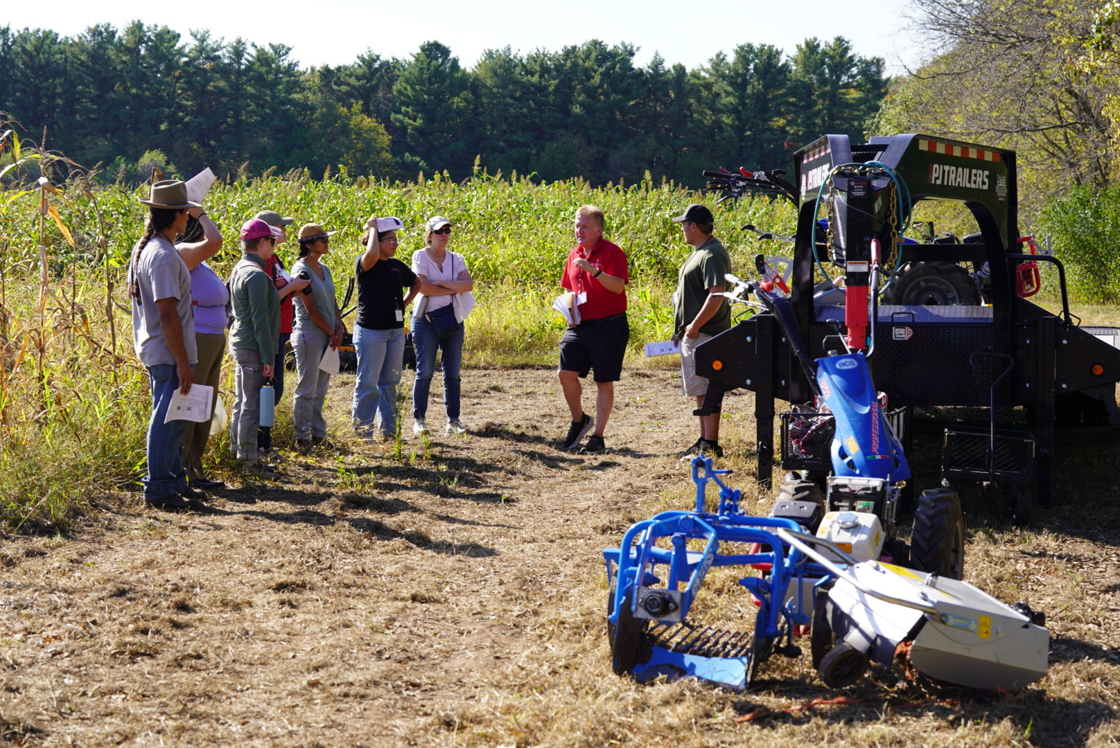 A group of people stand next to a trailer with a small walk behind tractor with a corn field in the background.