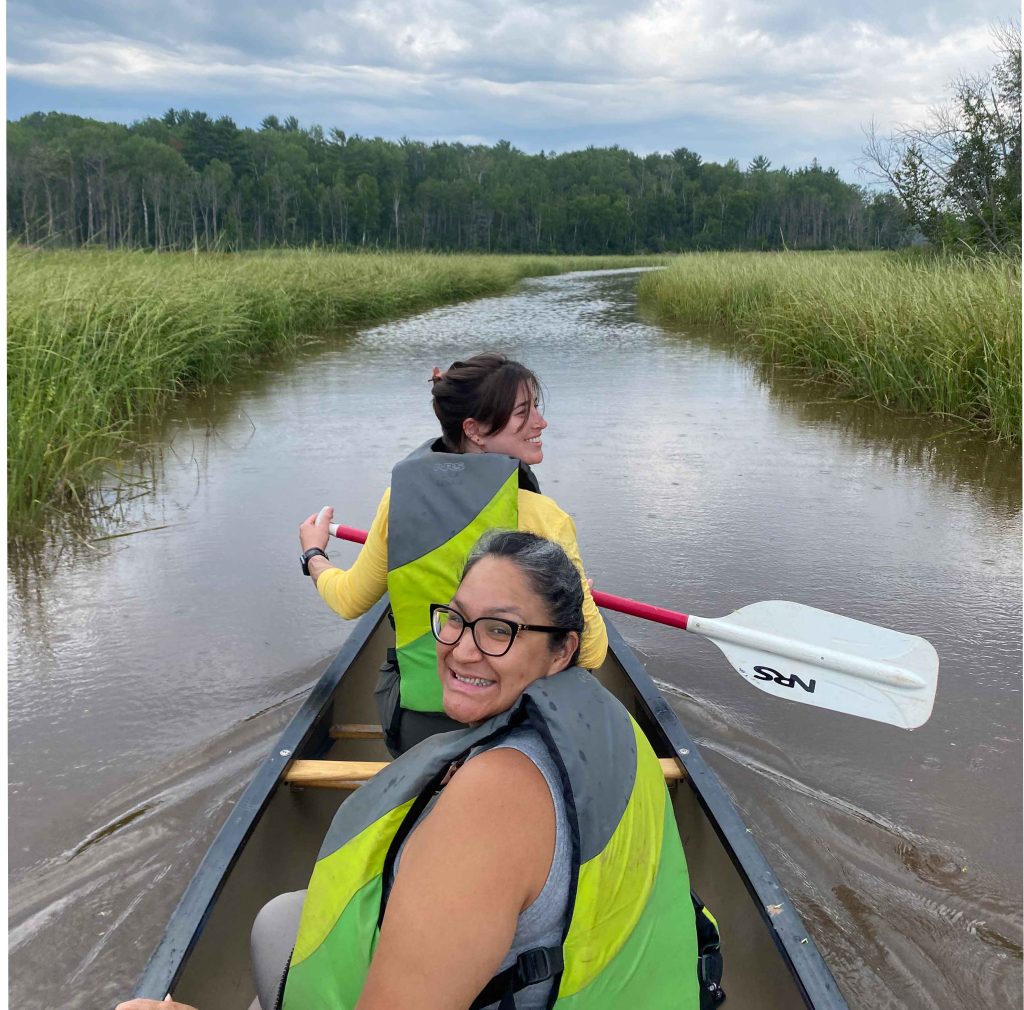 Becca Honeyball and Ashla Ojibwe smiling in a canoe surrounded by wild rice.