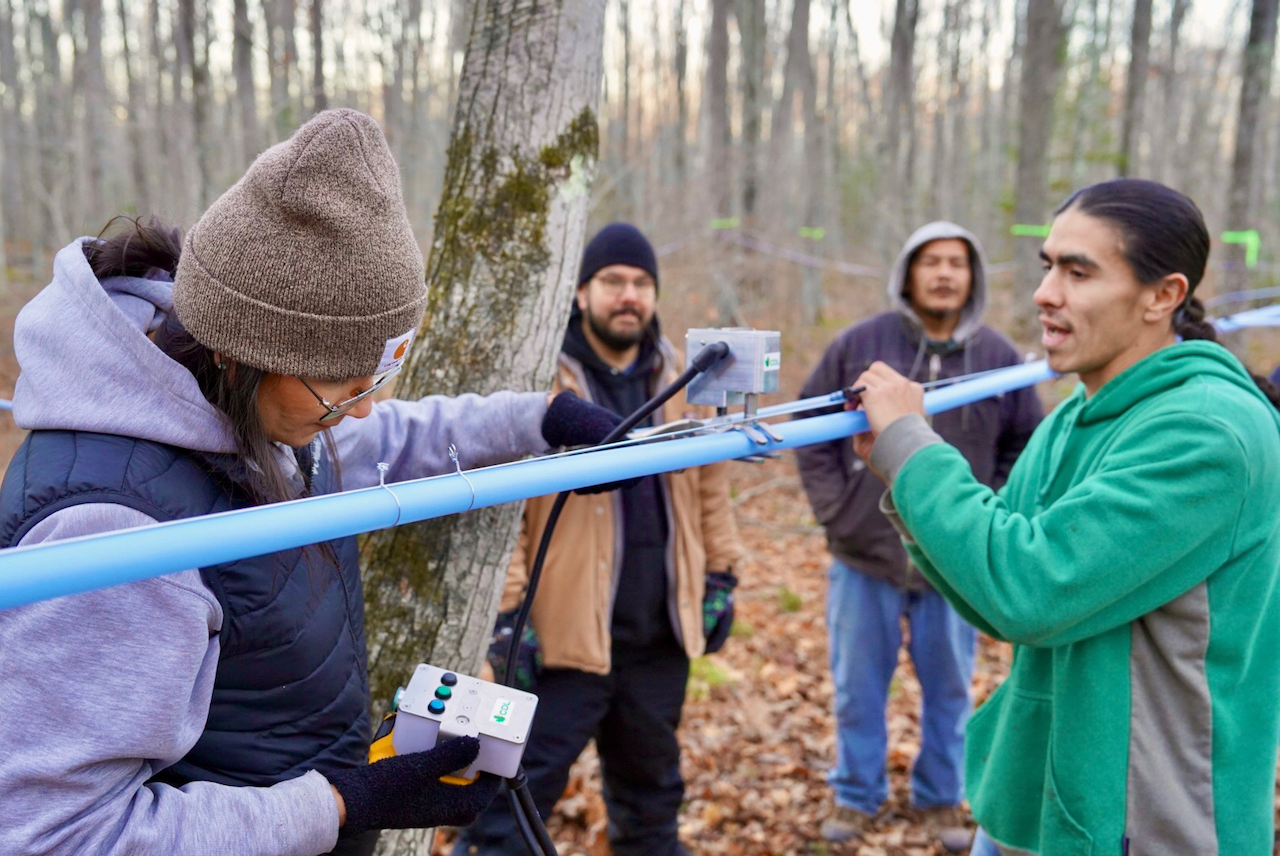 Closeup of 4 people holding onto a blue plastic maple sap tubing line with instructor explaining how to puncture the line to attach lateral lines.